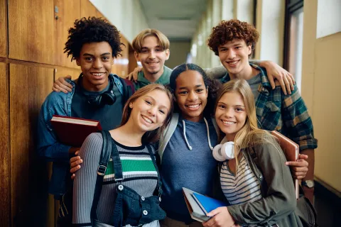 A group of 6 diverse teens holding backpacks and books, huddled together in a school hallway, smiling at the camera.