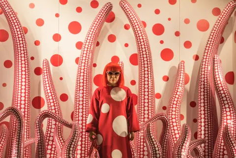 Japanese artist Yayoi Kusama in front of a white wall with red polka dots and surrounded by artwork of white tentacles with red polka dots